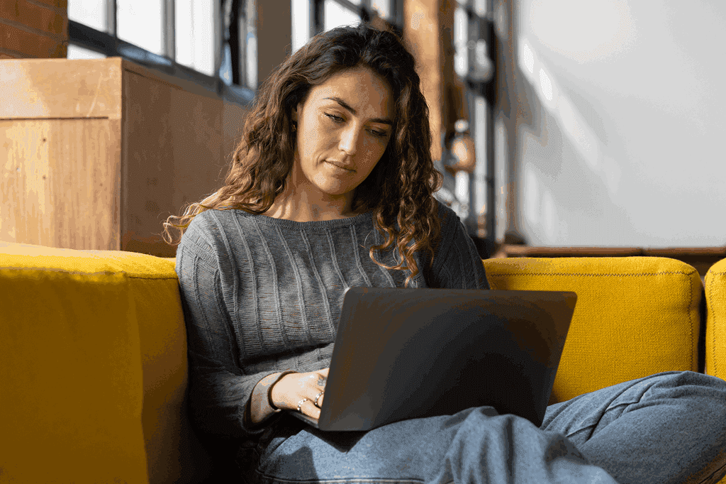 Mulher de cerca de 40 anos, de cabelos escuros cacheados e pele clara, sentada em um sofá, mexendo no notebook que está em seu colo, representando a ativação das APIs do C6 Bank.