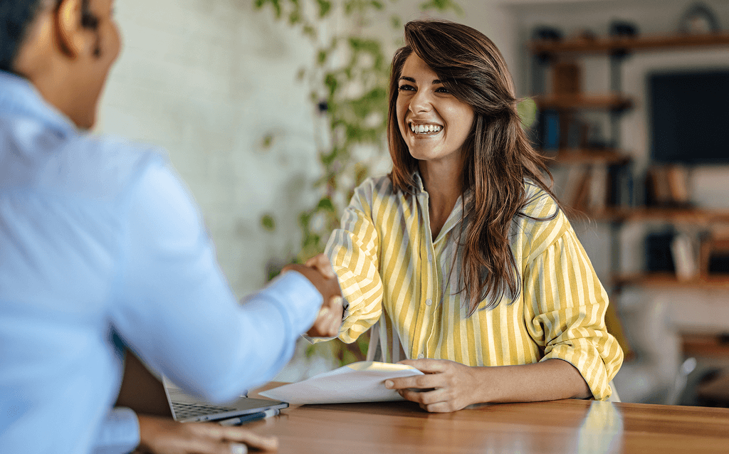 Mulher sorridente de camisa listrada amarela e branca aperta a mão de outra pessoa durante uma entrevista de emprego ou reunião profissional.