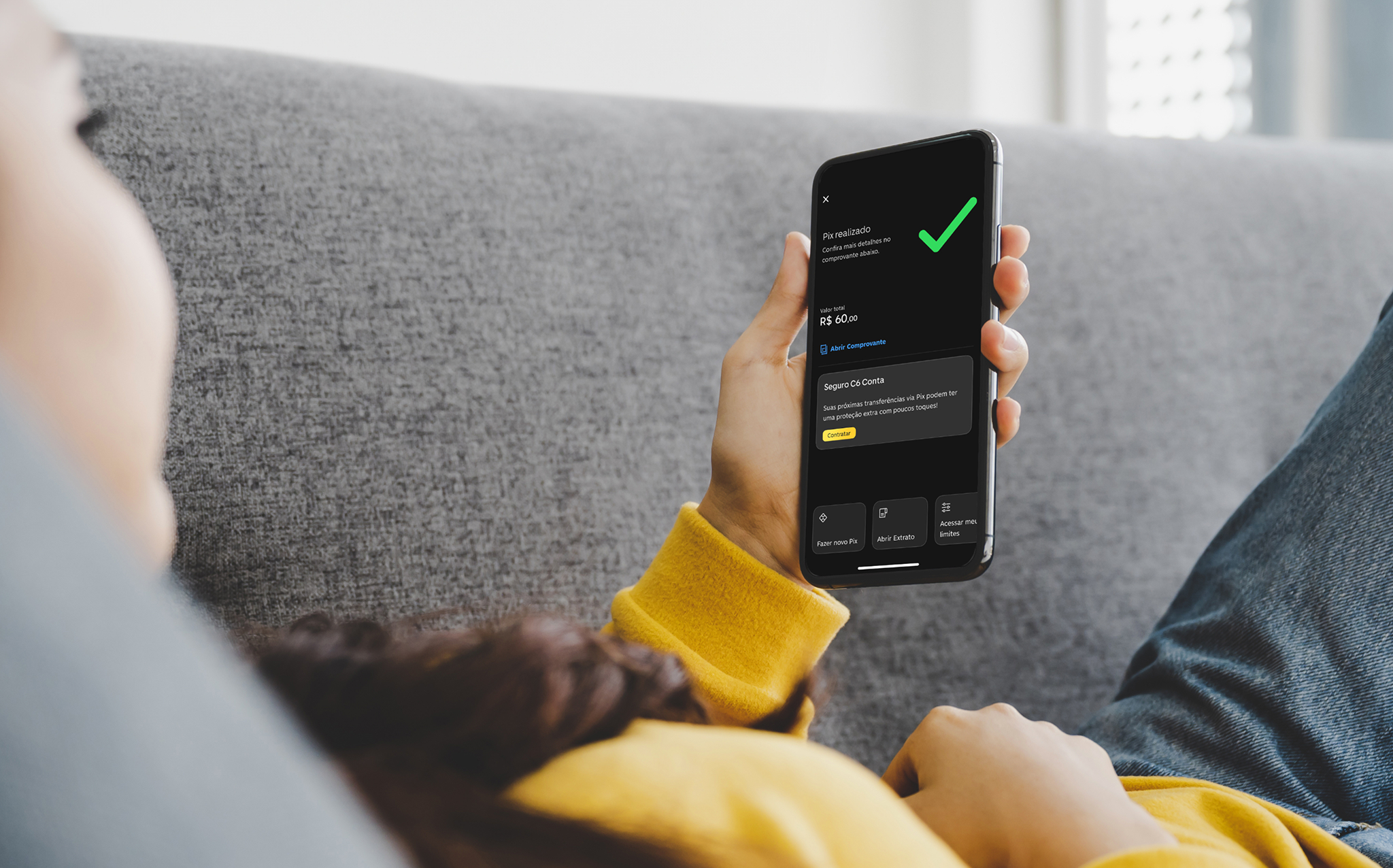 Pessoa deitada em um sofá, vestindo moletom amarelo, segurando um smartphone com a tela exibindo uma confirmação de Pix realizado no aplicativo do C6 Bank no valor de R$ 60,00.