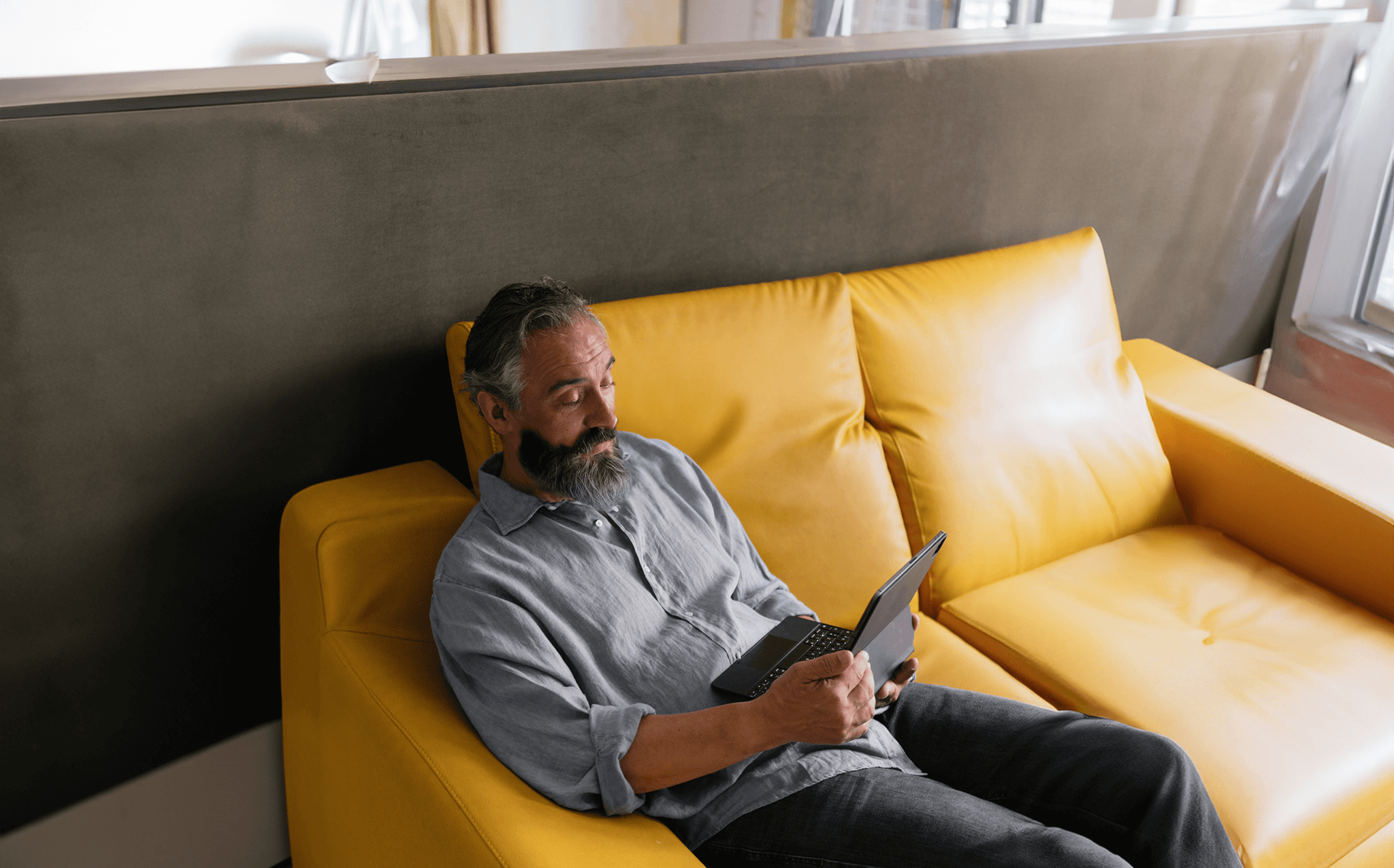 Homem de meia-idade com barba e cabelos grisalhos sentado em um sofá amarelo, usando um tablet com teclado. Ele está relaxado, vestindo uma camisa azul clara de linho e calça jeans, em um ambiente interno iluminado por luz natural.