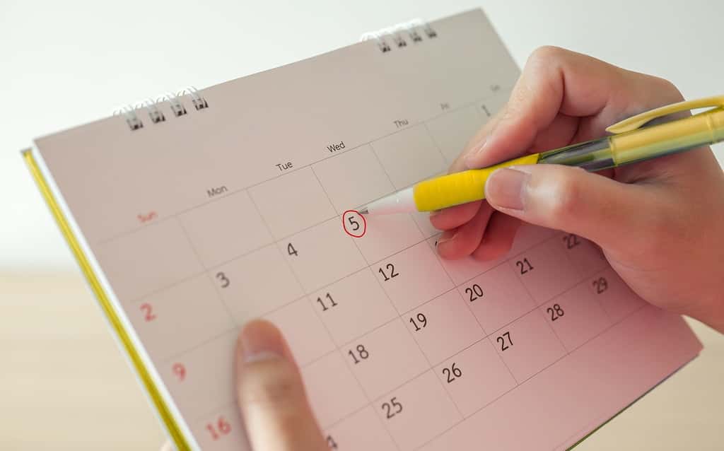 Pessoa segurando um calendário e circulando a data 5 com uma caneta amarela, simbolizando o agendamento ou controle de prazos importantes.