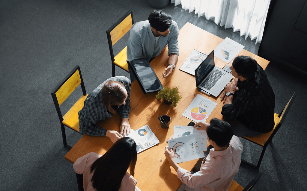 Cinco pessoas reunidas em torno de uma mesa de madeira em um ambiente de escritório, analisando gráficos e dados em papéis e laptops durante uma reunião.