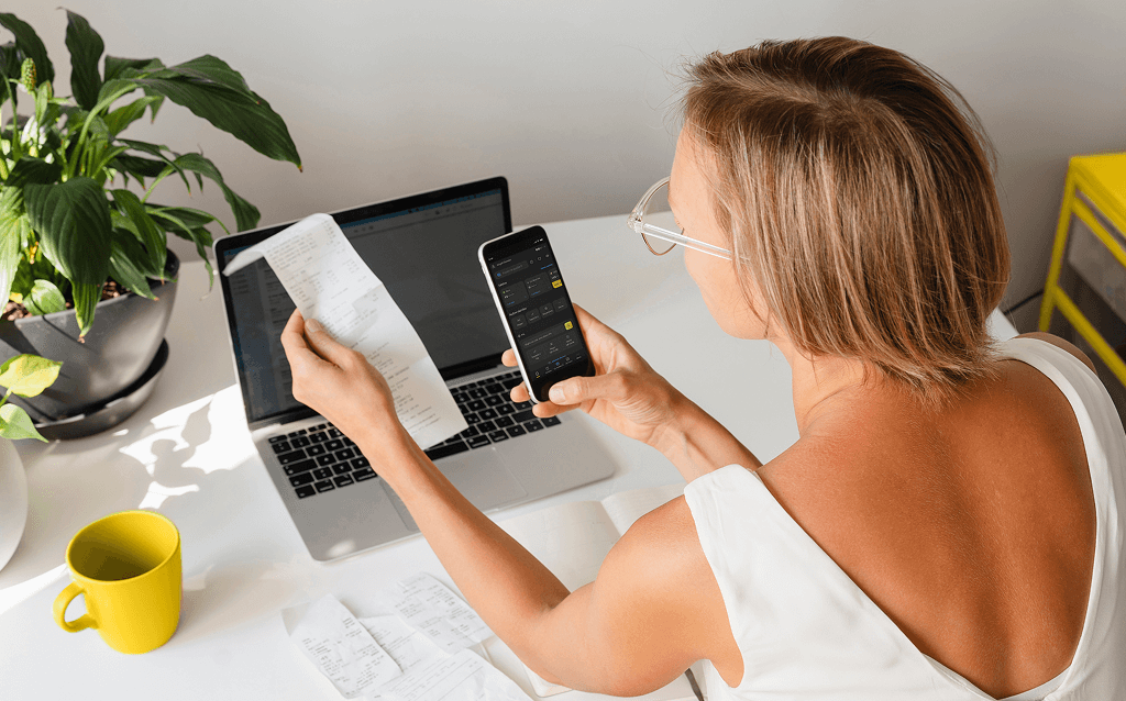 Mulher analisando recibos de compras enquanto confere informações financeiras no celular, com notebook aberto à sua frente sobre a mesa, ao lado de uma caneca amarela e uma planta.