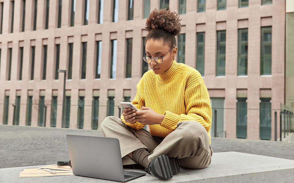 Pessoa sentada ao ar livre em frente a um prédio, usando suéter amarelo e calça bege, olhando para o celular com um notebook aberto à sua frente.