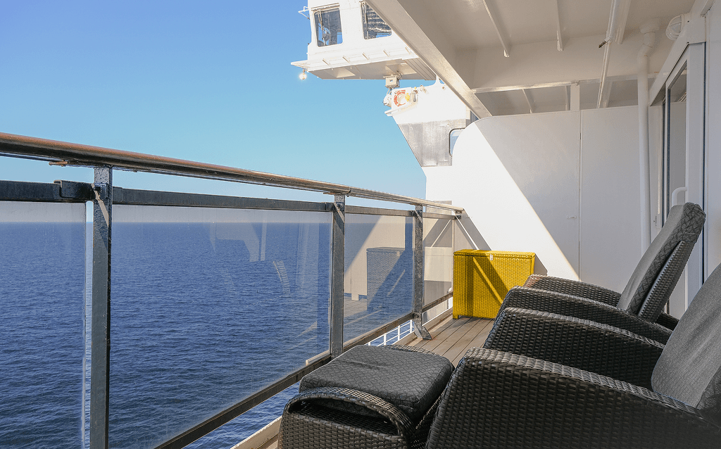 Varanda de cabine de cruzeiro com duas poltronas de vime, um banco amarelo e vista para o mar.
