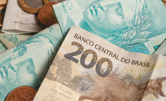 Cédula de 200 reais em destaque, cercada por notas de 100 reais e moedas do real brasileiro.