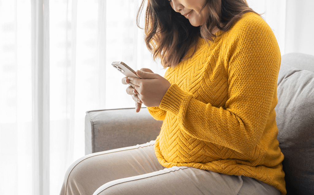 Mulher grávida de blusa amarela sentada no sofá utilizando o celular