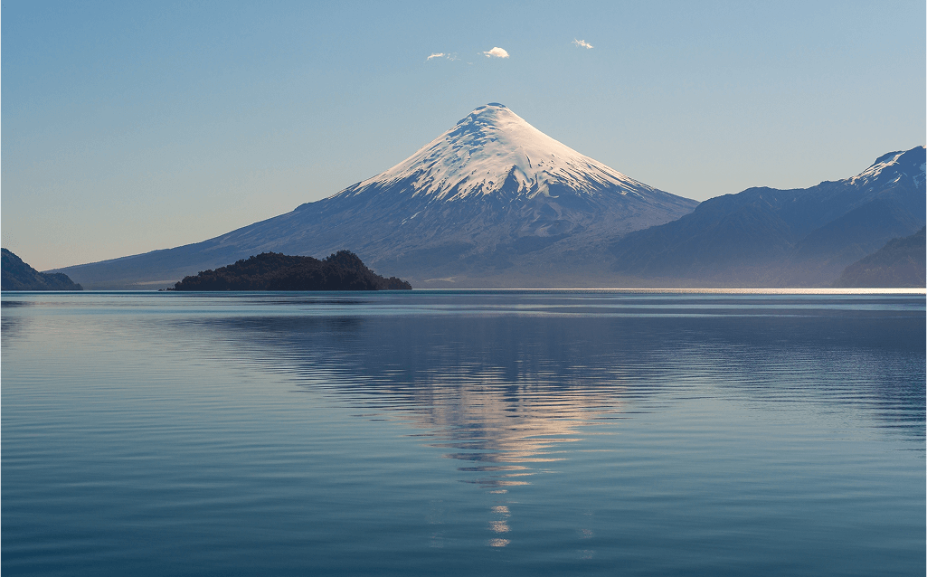Montanha com pico nevado refletida em um lago calmo, sob céu azul claro. A paisagem inclui ilhas e cordilheiras ao fundo.