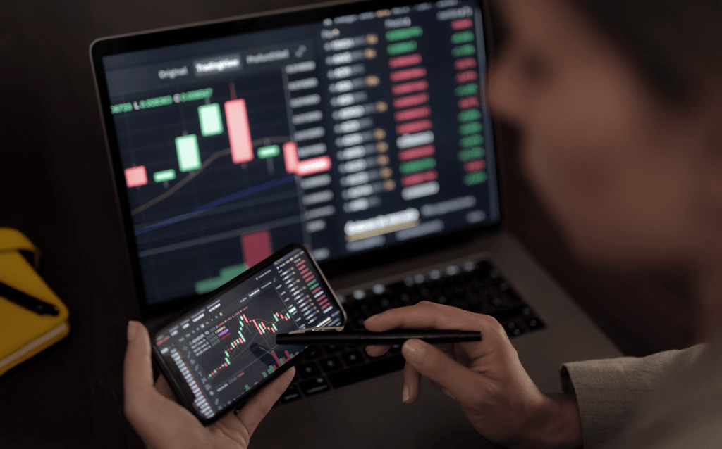 Pessoa olhando gráficos financeiros na tela do computador e smartphone, destacando o ambiente de investimento e representando a taxa de liquidação