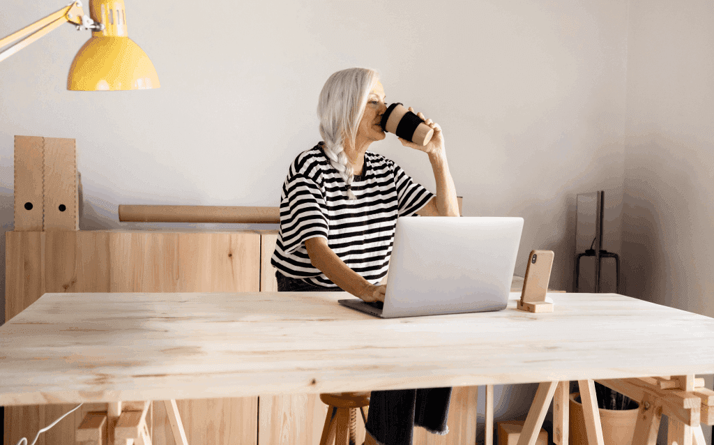 Mulher branca de cabelos grisalhos trabalhando como autônoma em um computador enquanto toma um café.