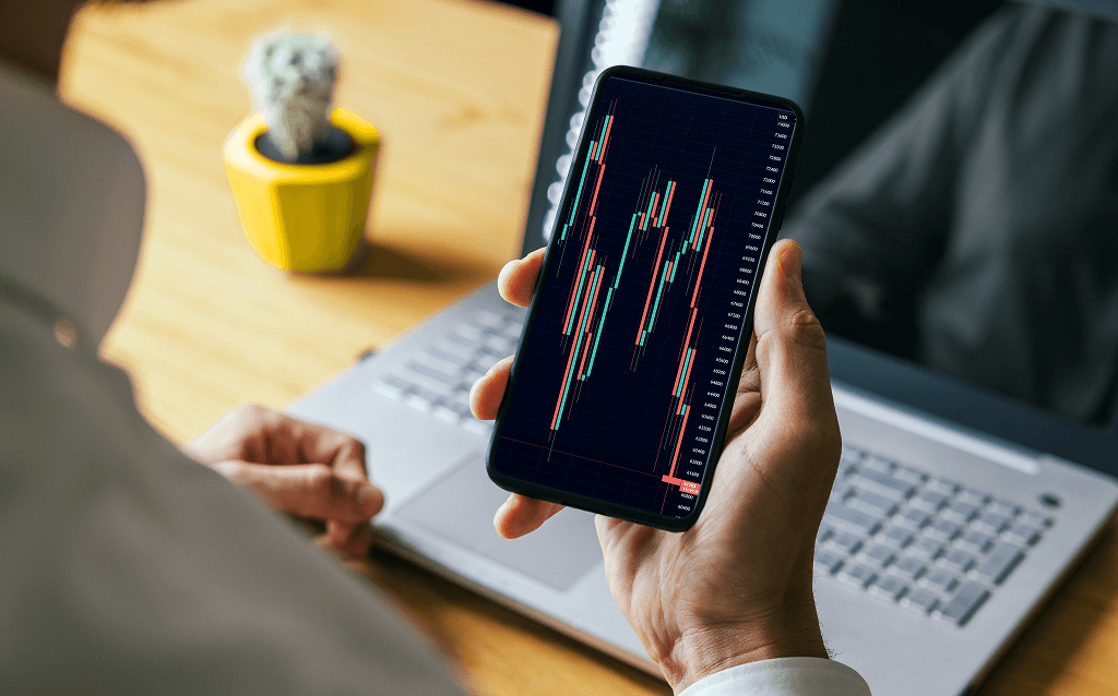 Pessoa segurando um smartphone que exibe um gráfico de velas (candlestick) com variações de preço, representando movimentações no mercado financeiro. Ao fundo, há um notebook e um vaso com um pequeno cacto sobre uma mesa de madeira, em ambiente de trabalho.