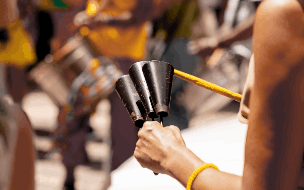 Mão de uma pessoa segurando um agogô de metal com três sinos pretos, tocado com uma baqueta amarela. A pessoa usa uma pulseira amarela no pulso. Ao fundo, aparecem outras pessoas desfocadas vestidas com roupas amarelas e instrumentos de percussão, sugerindo um ensaio ou apresentação de carnaval ao ar livre, com foco na cultura brasileira e na música percussiva.