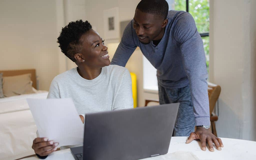 No centro da composição, uma mulher sentada segura uma folha de papel na mão esquerda enquanto olha para um homem que está inclinado ao lado dela, apoiando as mãos sobre a mesa. À frente deles, há um notebook aberto sobre a mesa branca. O ambiente é interno e residencial, com cama ao fundo, janela lateral com luz natural e decoração discreta.