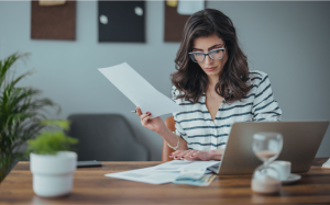 Mulher jovem, branca, de cabelos castanhos, blusa listrada azul e branca, e óculos, sentada em uma escrivaninha de madeira com diversos papéis e um notebook, segurando um dos papéis em mãos, direcionando o olhar para os da mesa, representando alguém começando a investir.