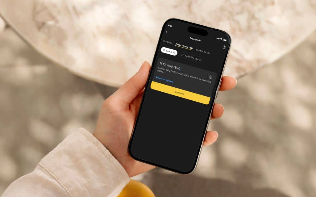 Pessoa segurando um smartphone com a tela de transferência Pix aberta no aplicativo da Conta C6, exibindo campo de chave Pix, valor e botão de continuar em fundo escuro.