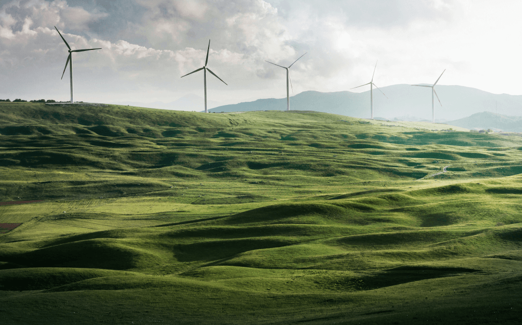 Paisagem de colinas verdes onduladas com vários aerogeradores (turbinas eólicas) distribuídos ao fundo, sob um céu parcialmente nublado, representando geração de energia limpa e sustentável.