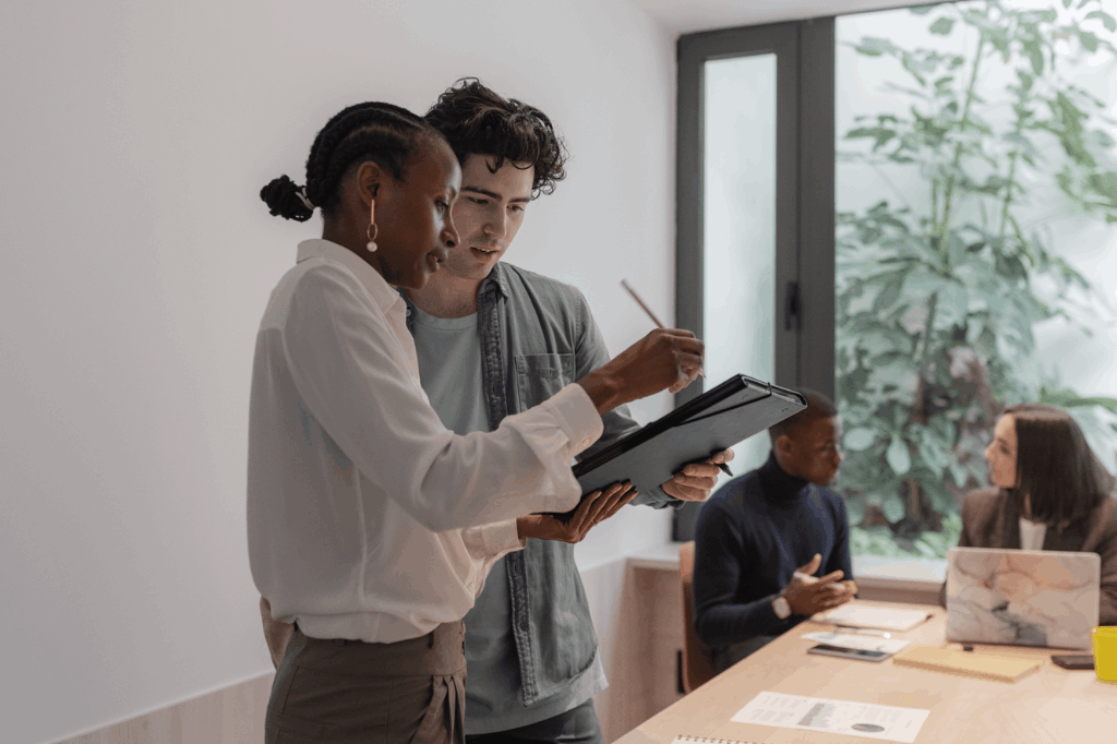 Uma mulher negra, de cabelos presos e roupa social, apresenta em um tablet para um jovem branco, de cabelos pretos e roupa casual, algo importante em um tablet. Ambos estão em pé, em um ambiente profissional, representando uma conversa sobre empréstimo consignado clt, com pessoas sentadas atrás discutindo em um contexto empresarial.