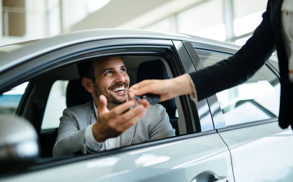 No centro da composição, um homem sentado dentro de um carro branco, com a janela aberta, estende a mão para receber uma chave automotiva de outra pessoa posicionada do lado de fora do veículo. O homem está sorrindo e veste blazer claro. A outra pessoa aparece parcialmente na lateral direita da imagem, com o braço estendido segurando a chave. O ambiente é interno, bem iluminado, com grandes janelas ao fundo, caracterizando uma concessionária ou showroom de veículos.