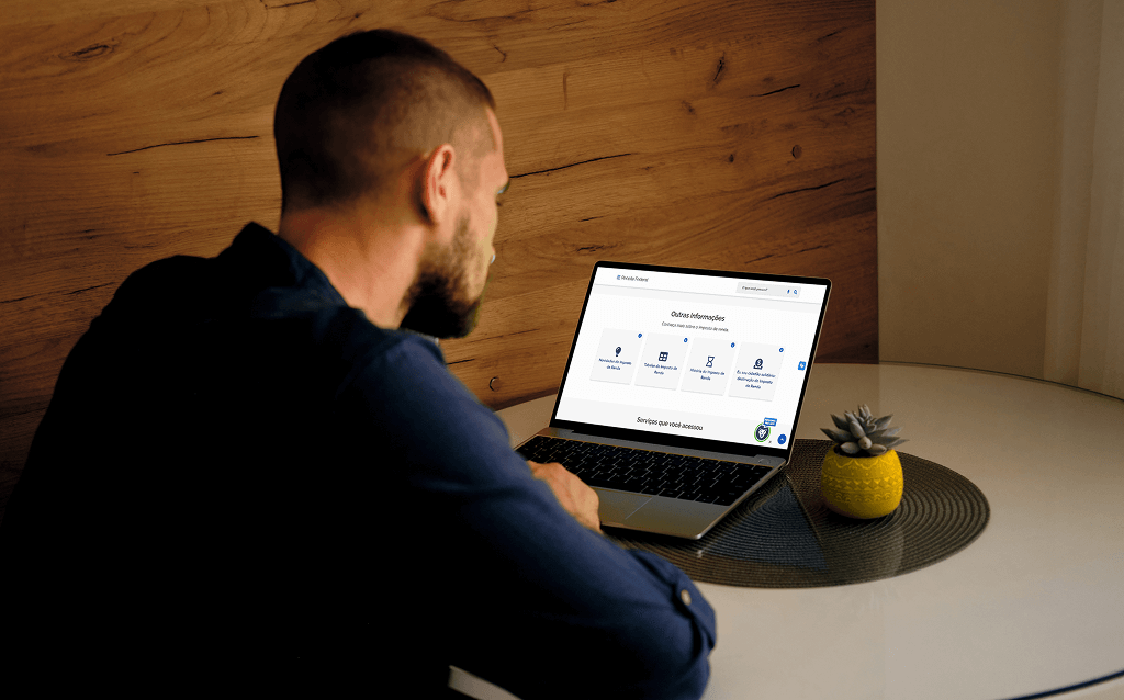 Homem de barba, sentado à mesa redonda com toalha cinza, usando um notebook que exibe a página da Receita Federal do Brasil. Ao lado do computador, há um vaso amarelo com uma suculenta.