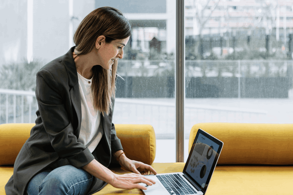 Mulher loira sentada em um sofá amarelo, mexendo no notebook, representando o uso de um gerenciador financeiro. Ao fundo, um cenário urbano, com prédios corporativos.