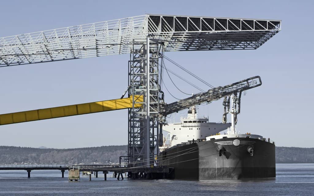 Navio cargueiro atracado em terminal portuário, com estrutura metálica e esteiras de carregamento usadas em operações de importação e exportação de mercadorias.