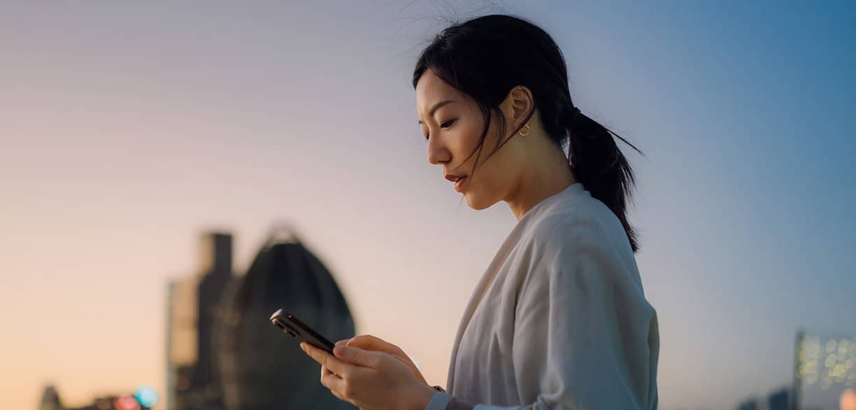 Mulher olhando o celular ao ar livre durante o entardecer, com prédios ao fundo desfocados, representando acompanhamento de resultados financeiros pelo smartphone.
