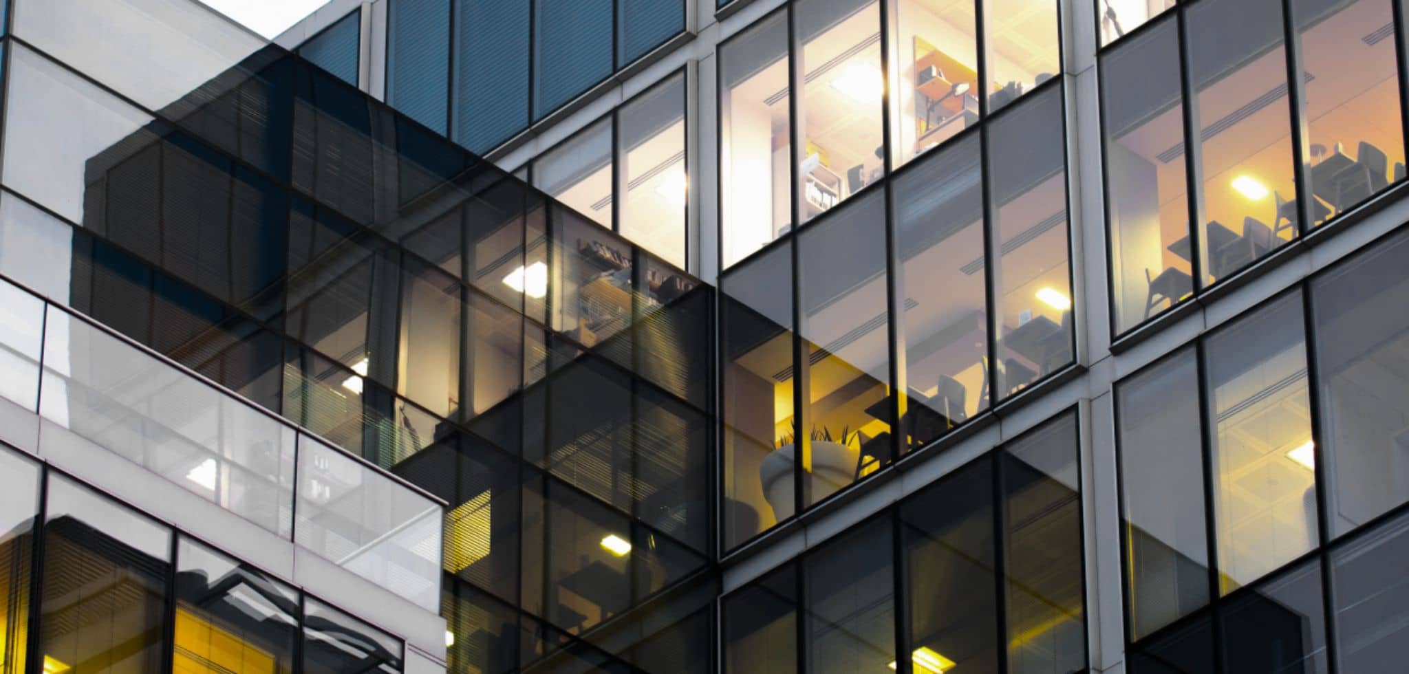 A composição mostra a parte externa de um prédio corporativo moderno com fachada de vidro e estrutura metálica. É possível ver, através das janelas, salas internas iluminadas com mesas, cadeiras e objetos de escritório. A perspectiva é inclinada, destacando a arquitetura contemporânea e o ambiente empresarial.