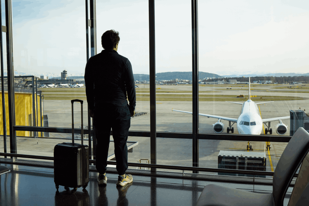 Homem de costas parado em um andar alto de um aeroporto, olhando o avião pousado do lado de fora, representando a vida de um nômade digital.