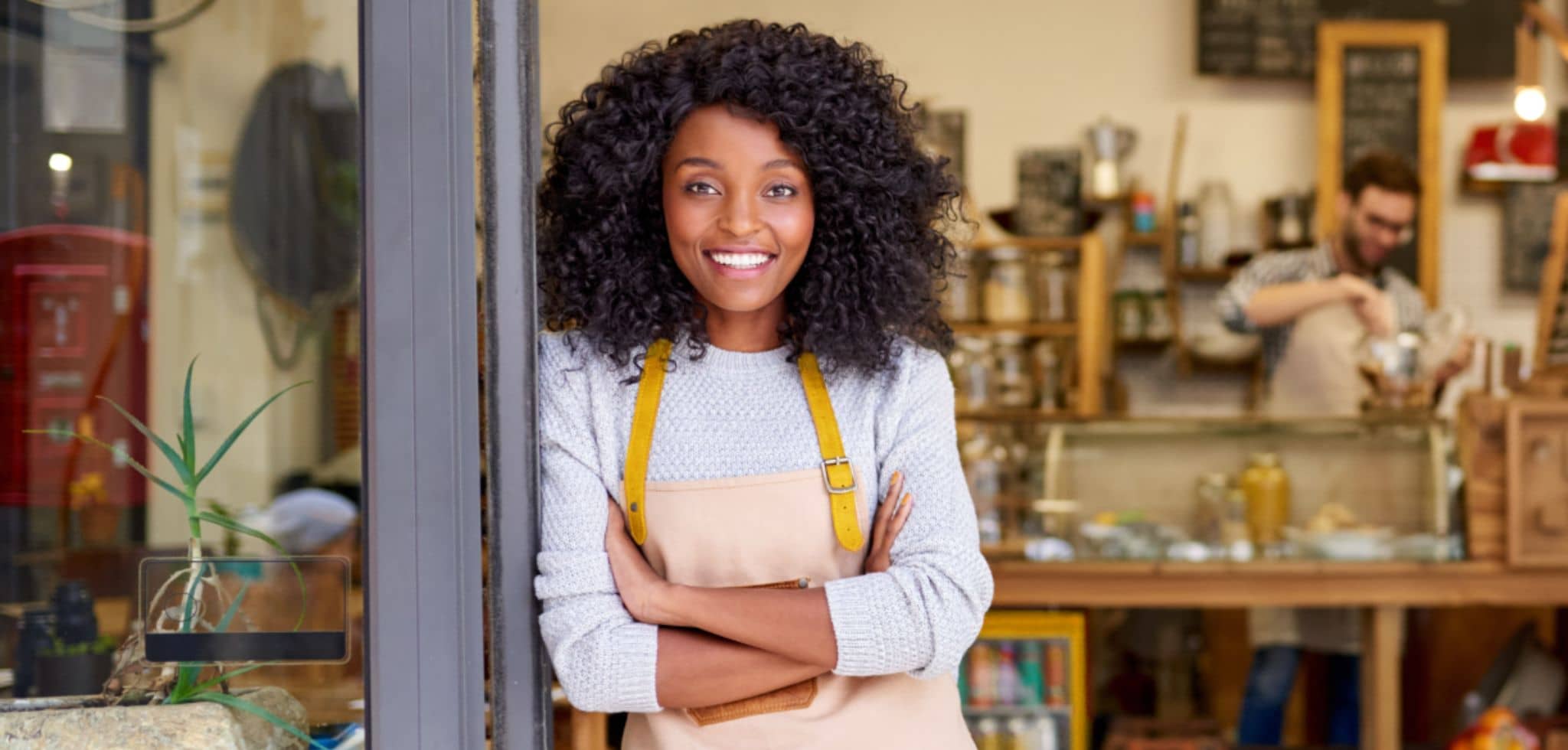 No centro da composição, uma mulher está posicionada na entrada de um estabelecimento, com os braços cruzados e sorriso voltado para a câmera. Ela veste blusa clara e avental bege com alças amarelas. Ao fundo, dentro do estabelecimento, há balcão com produtos e um funcionário trabalhando atrás do caixa. O ambiente é interno, com prateleiras, utensílios e elementos típicos de cafeteria.