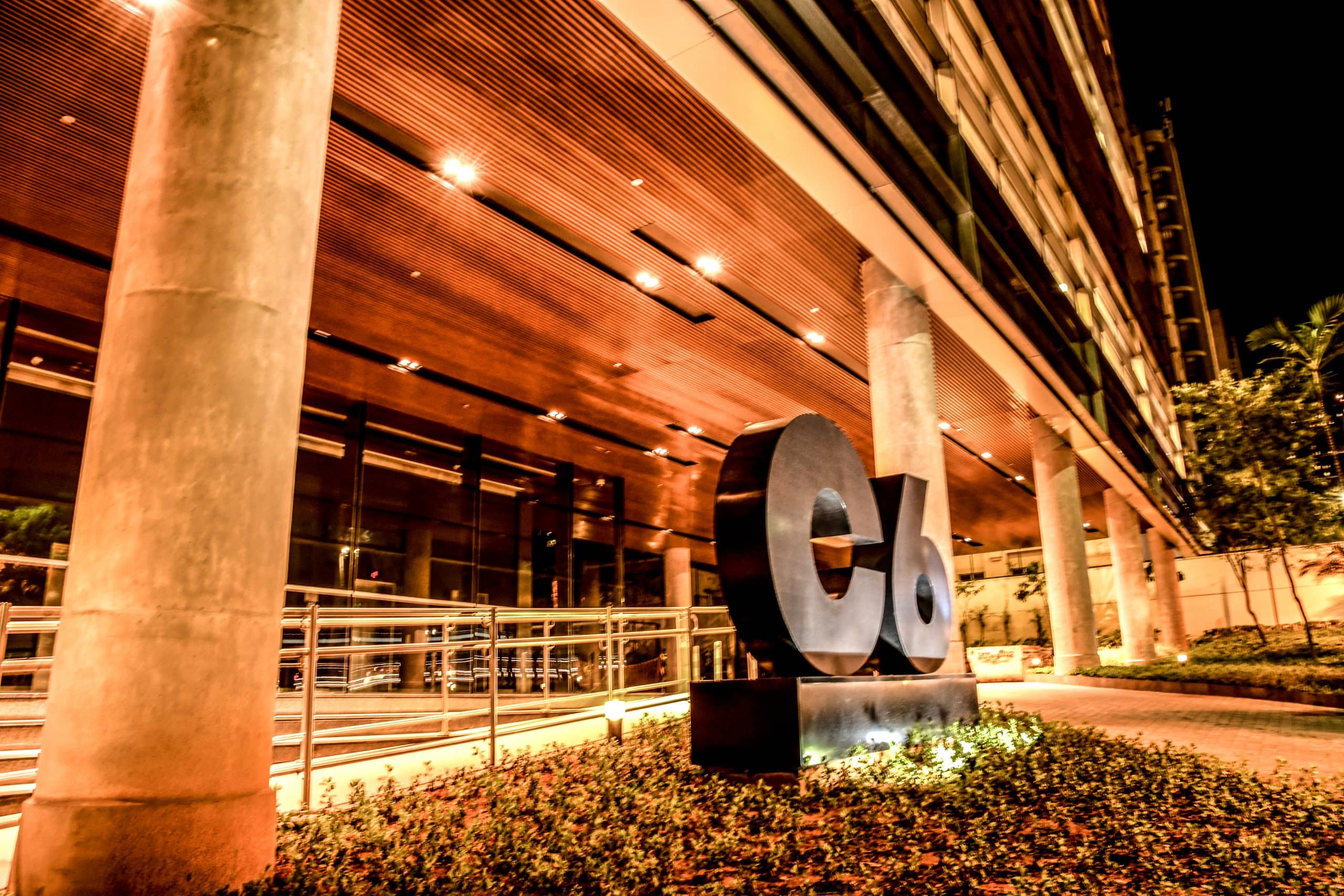 Foto da fachada do prédio do C6 Bank, onde se visualiza o logo do C6 Bank em frente ao prédio em meio ao jardim, com as luzes do teto da recepção em tons de amarelo em destaque.