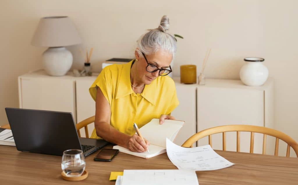 Mulher madura sentada à mesa organizando documentos e anotando informações em um caderno, com laptop e celular ao lado, representando planejamento financeiro na fase 60+
