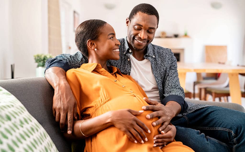 casal de mulher e homem negros sorri enquanto pensa em como montar uma poupança para filhos. a mulher está grávida e olhando para o rosto do marido, que passa o braço por cima do ombro dela e apoia a outra mão na barriga da mulher