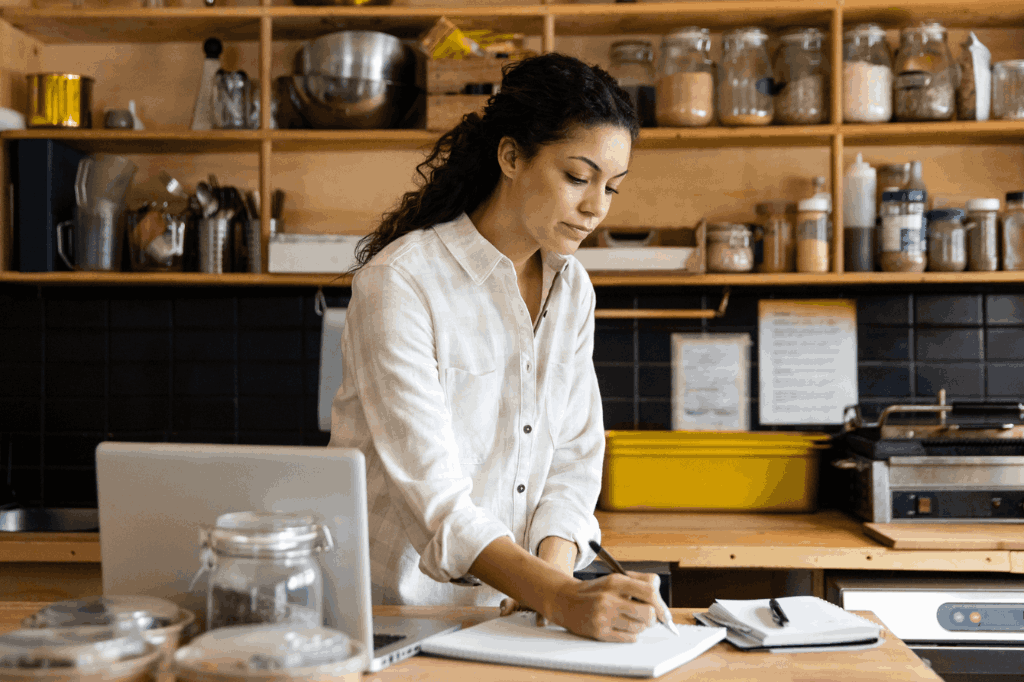 Empreendedora em sua própria cafeteria, anotando em um caderno com uma caneta, representando o estudo para escolher a razão social da sua empresa. Ela é uma mulher de cabelos pretos, cacheados e olha para o caderno com uma expressão séria.