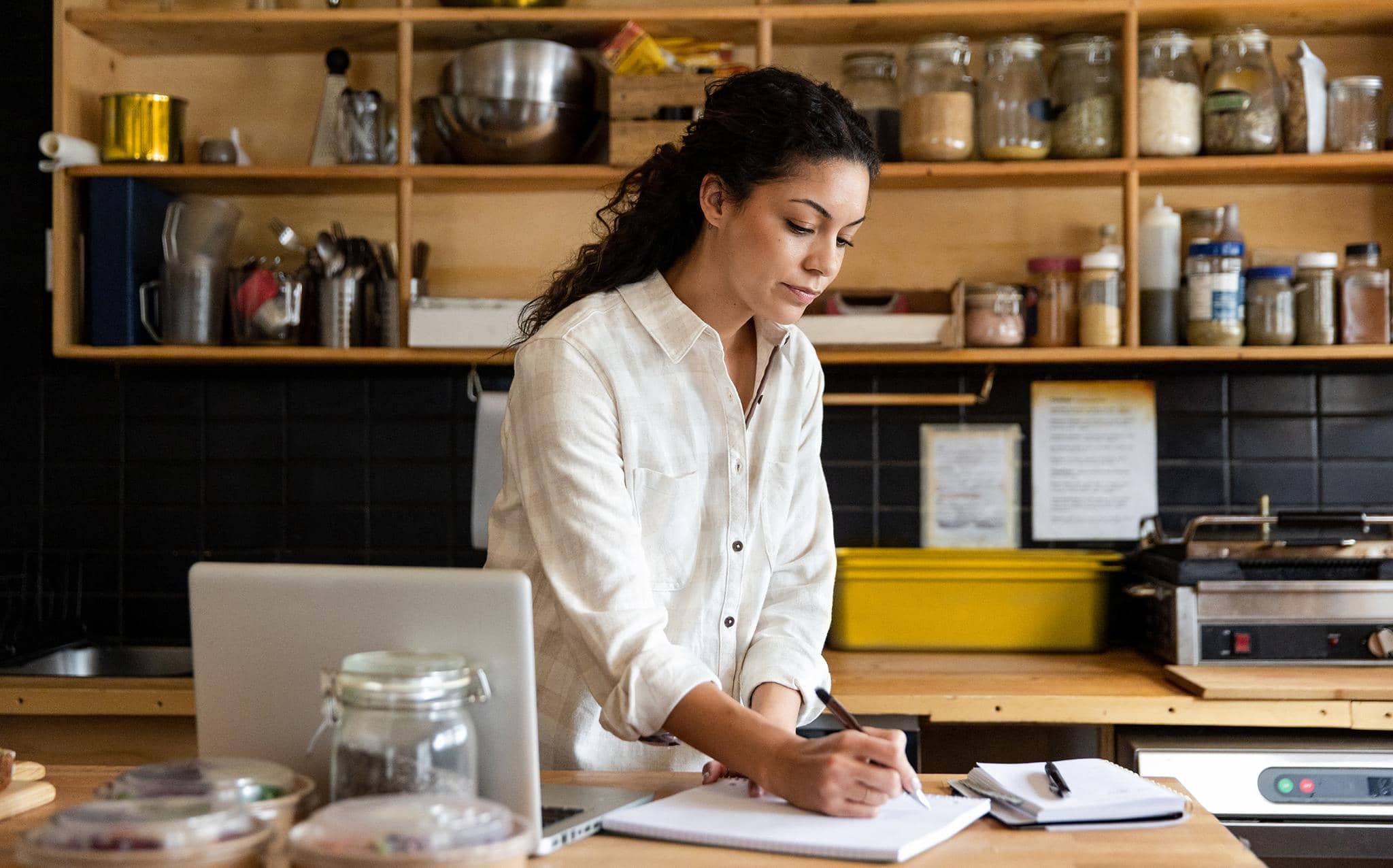 Mulher empreendedora em uma cozinha comercial fazendo anotações em um caderno, com notebook e potes de ingredientes ao redor, representando o controle financeiro de um pequeno negócio.