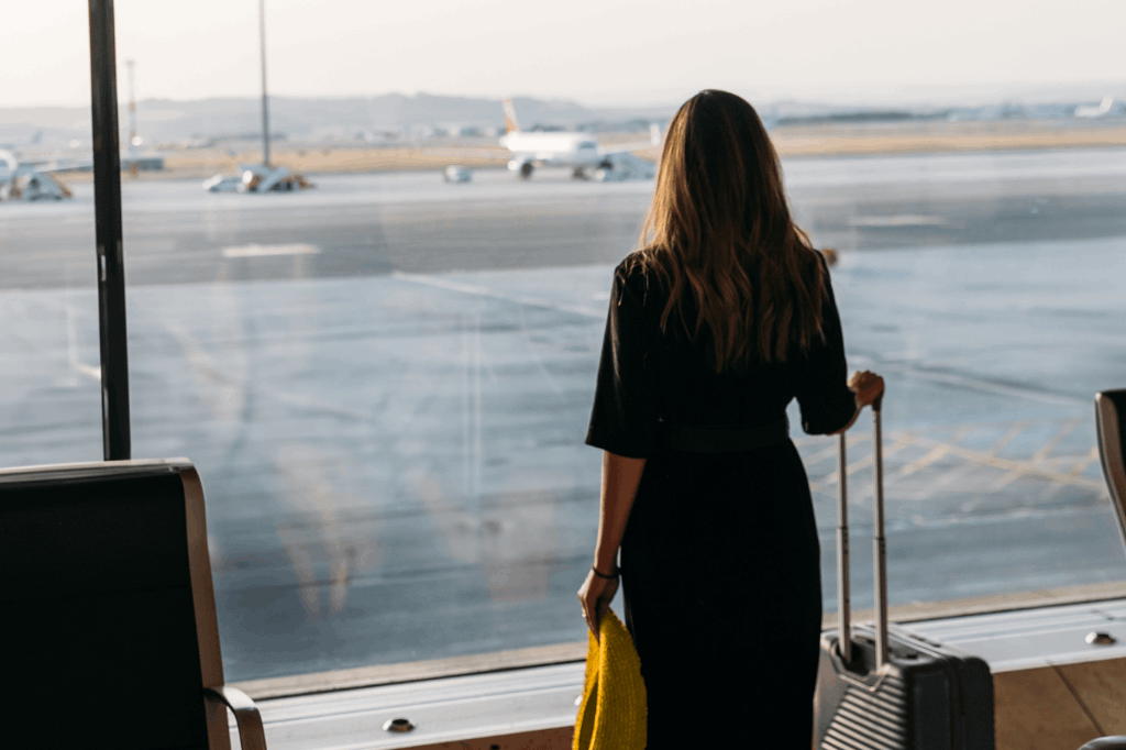 Mulher de costas para a foto em um aeroporto, com uma mala de rodinha em mãos, observando uma pista de aterrissagem em um aeroporto, representando o processo de renovar passaporte.