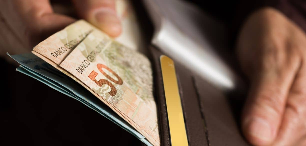 Pessoa retirando notas de cinquenta reais de uma carteira marrom, com cartão dourado visível no interior.