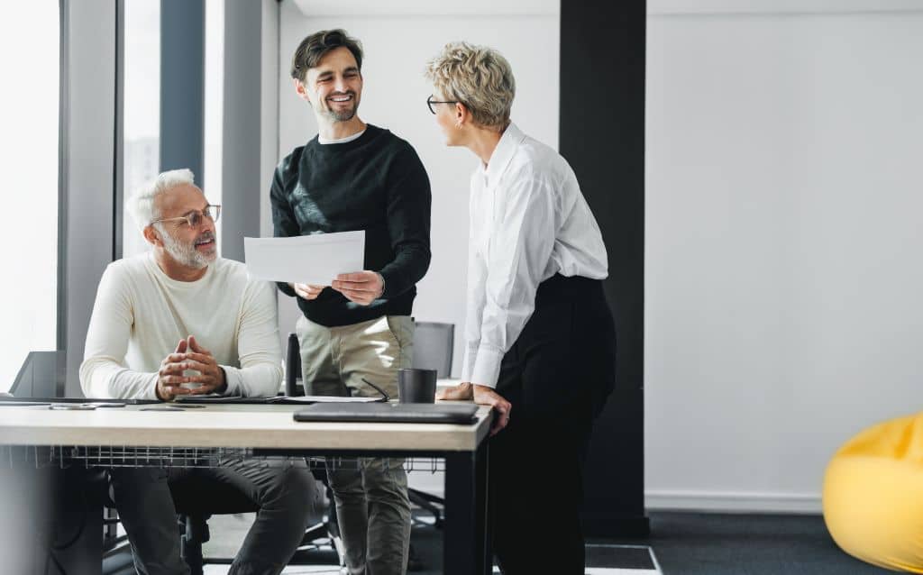 No centro da composição, aparece um homem em pé segurando uma folha de papel branca com as duas mãos, olhando para uma mulher que está apoiada na mesa. À esquerda, um homem sentado observa a conversa com as mãos apoiadas sobre a mesa. Todos estão em um escritório moderno, com mesa clara, cadeira, notebook fechado, caneca e objetos de trabalho visíveis. Ao fundo, há paredes claras, janelas amplas e elementos arquitetônicos em tons neutros, compondo um ambiente corporativo e profissional.