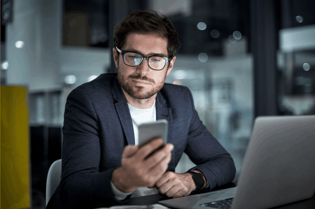 Homem branco empresário vestido socialmente segurando e olhando para um celular em suas mãos, representando o acompanhamento do ticket médio.