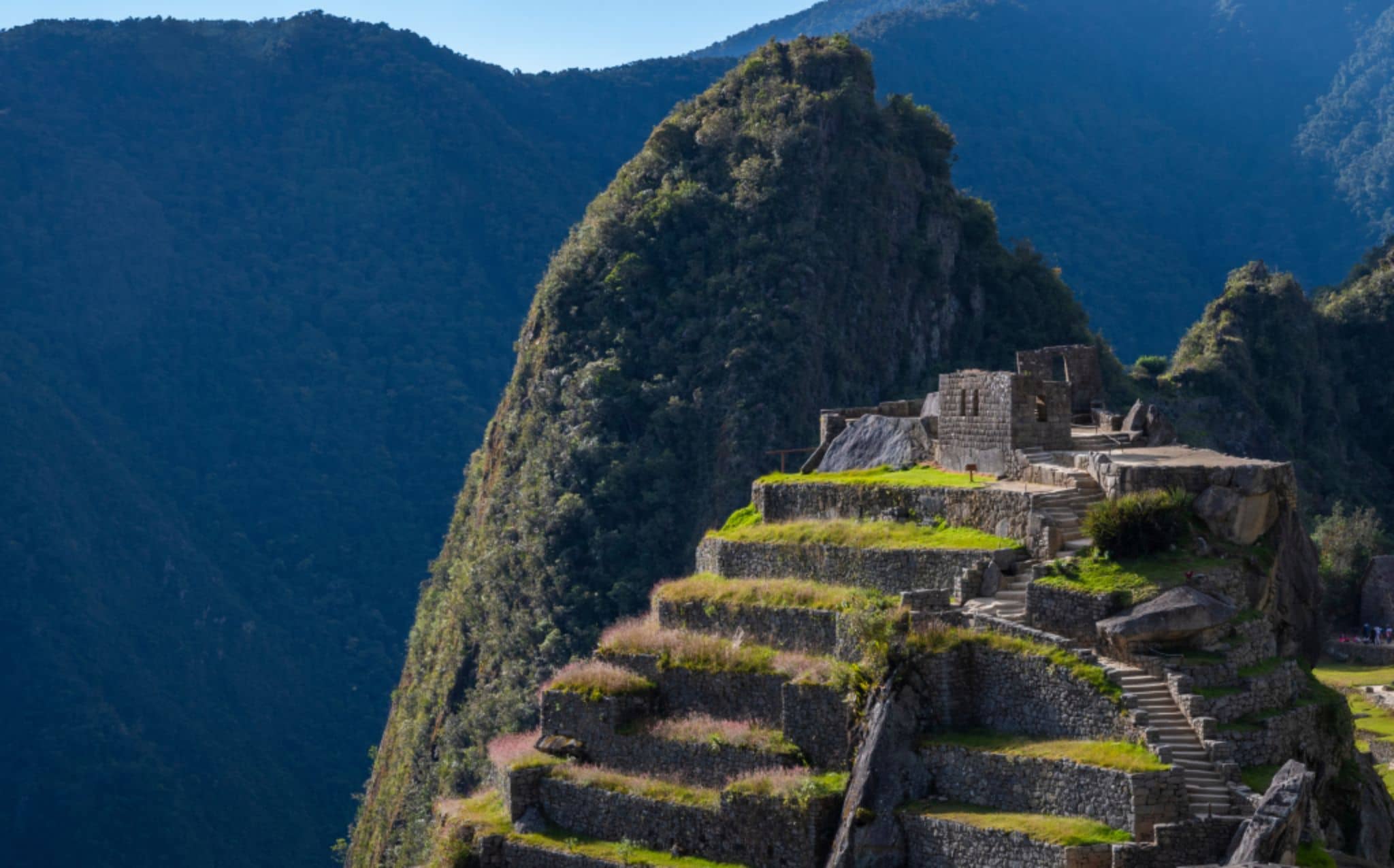 A imagem não apresenta textos visíveis. No centro da composição, aparecem estruturas de pedra e terraços escalonados localizados no topo de uma montanha. Ao fundo, há formações montanhosas cobertas por vegetação, compondo paisagem natural ampla. As construções estão distribuídas em níveis horizontais com escadarias de pedra conectando os diferentes patamares.