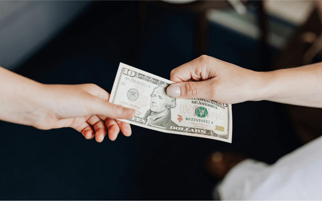 A mão de duas pessoas de pele clara segurando uma nota de 10 dólares que estampa o rosto de Alexander Hamilton, primeiro secretário do Tesouro dos EUA