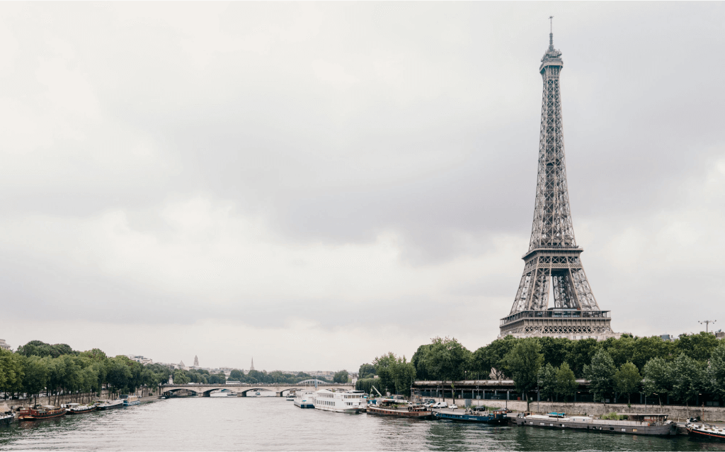 A Torre Eiffel está a direita da imagem. Barcos e árvores compõe a imagem junto com o Rio Sena.