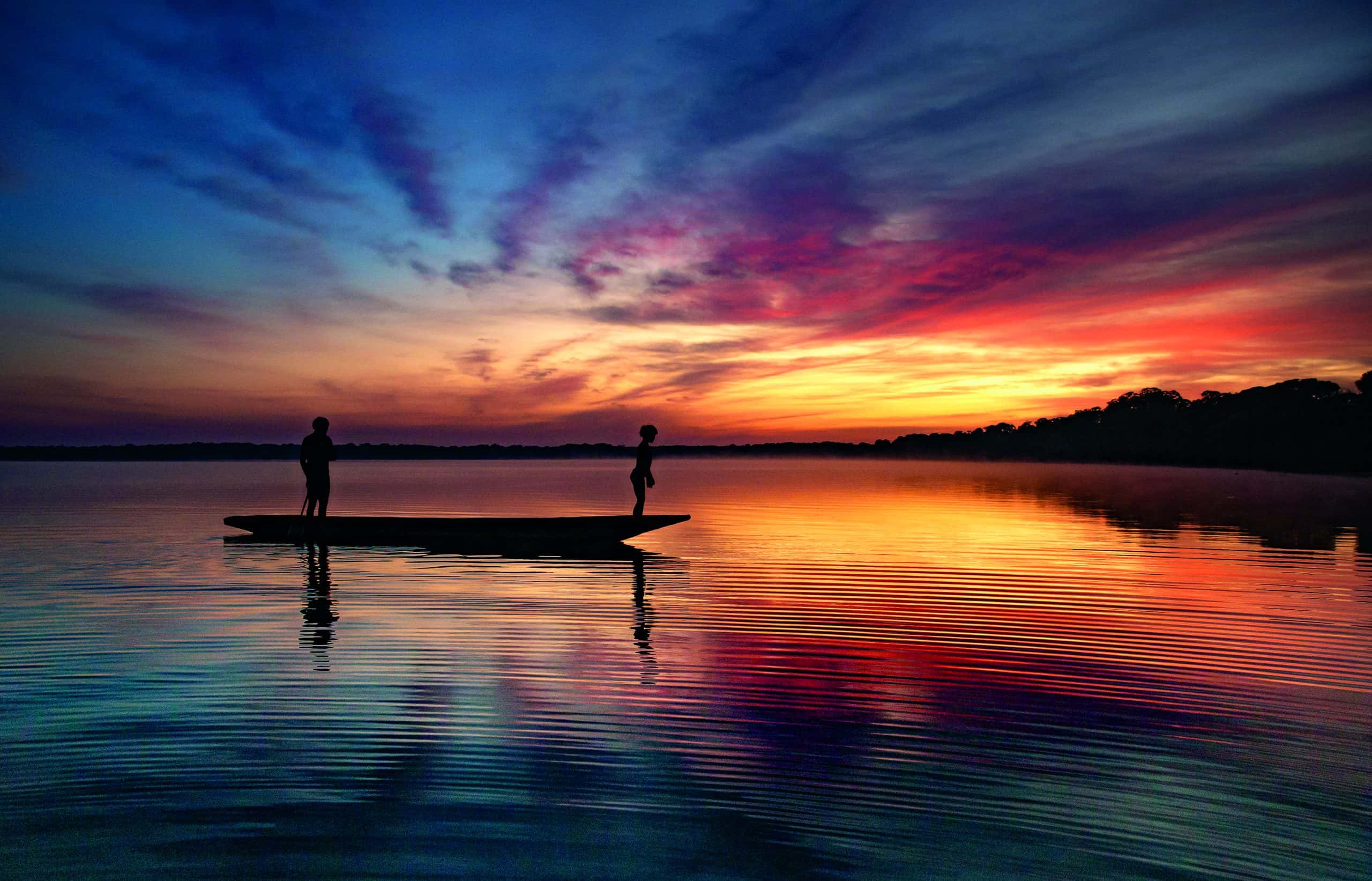 Foto de Araquém Alcantara de uma canoa no rio Amazonas onde se pode ver a silhueta de duas crianças em cima do barco e o pôr do sol ao fundo refletindo nas águas do rio.
