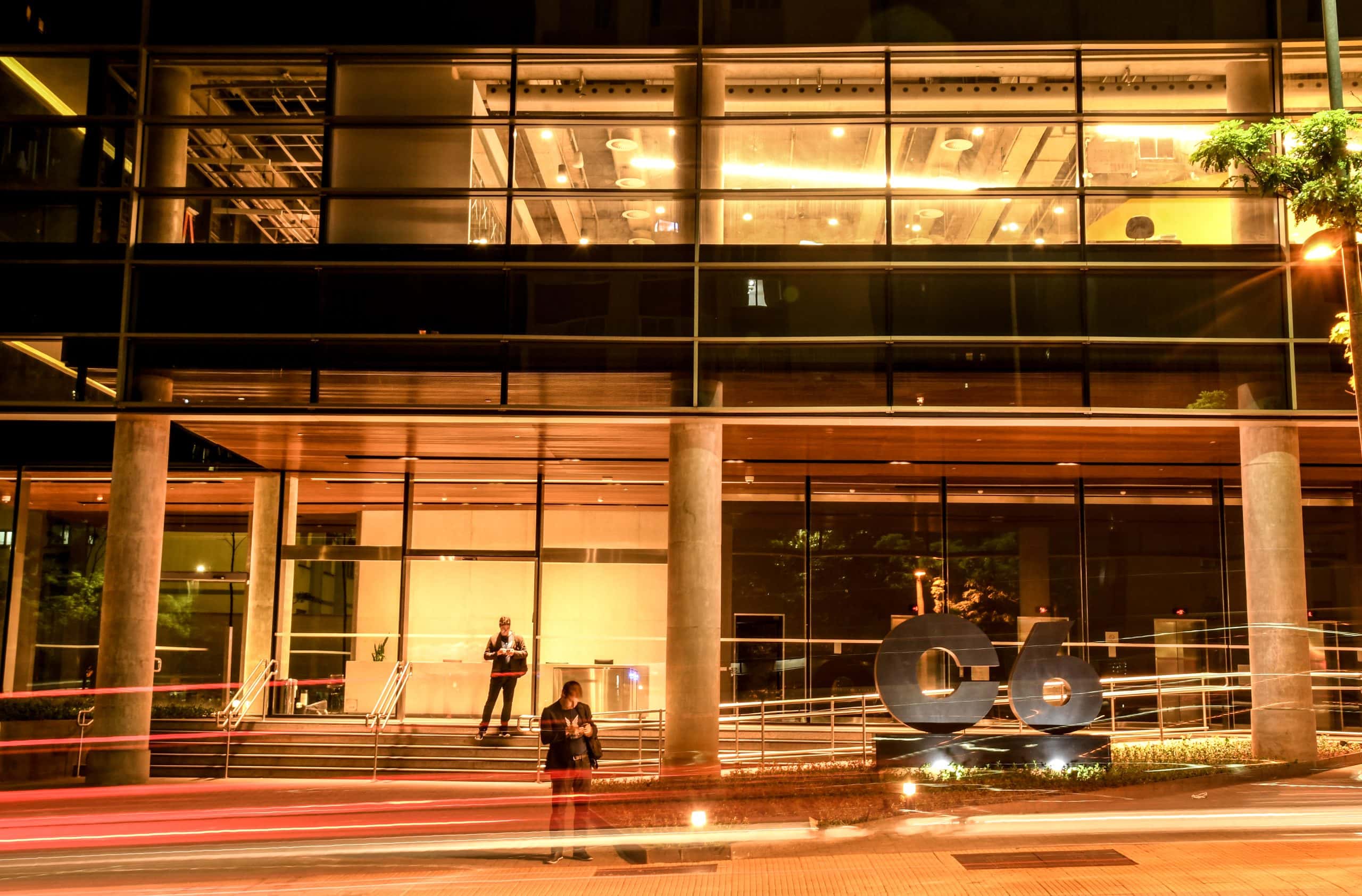 Foto noturna da fachada do prédio Carbon do C6 Bank, com o logo do C6 no jardim frontal do prédio em primeiro plano, a fachada do térreo iluminada com luzes em tom de bronze e duas pessoas ao fundo em pé