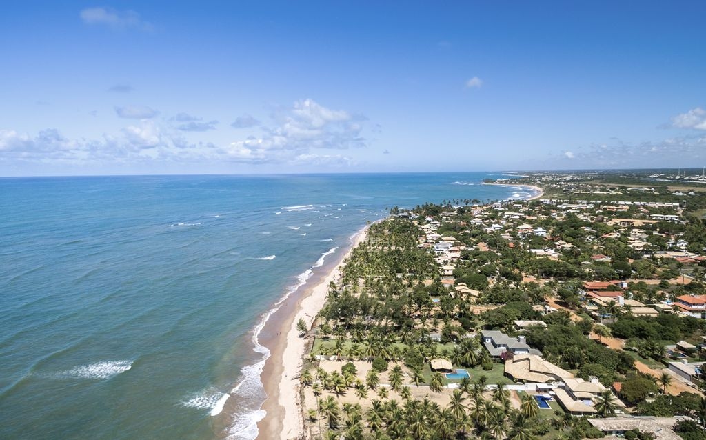 costa do sauipe praia, céu azul e depois da areia da praia é possível observar casas e residências luxuosas com vegetação comum em ambientes litorâneos