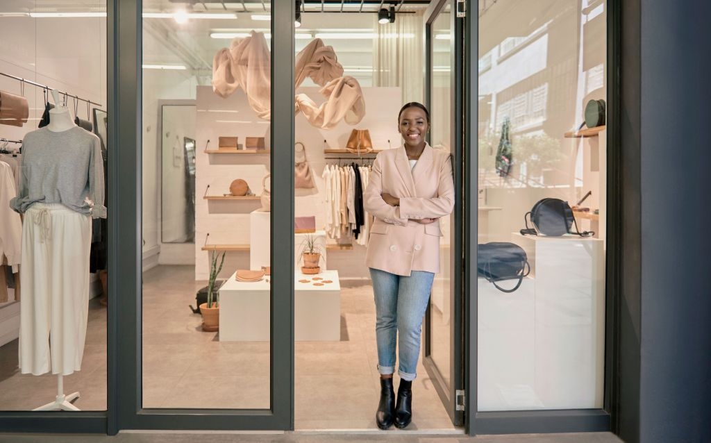 declaração anual mei -mulher negra vestindo calça jeans, botas pretas e casaco rosa elegante parada de braços cruzados e sorrindo em frente a uma loja