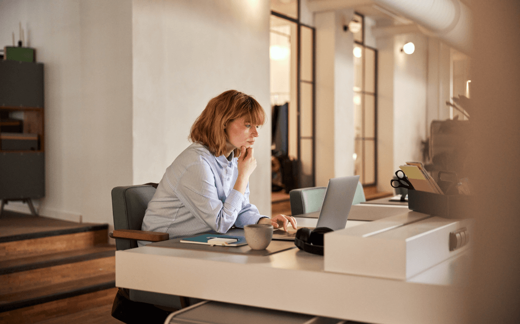 Mulher concentrada, sentada em um ambiente de trabalho moderno, olhando para a tela do notebook com foco. Sobre a mesa há uma caneca, bloco de anotações e itens de escritório. A imagem representa a importância da educação financeira como ferramenta de empoderamento e tomada de decisões conscientes.