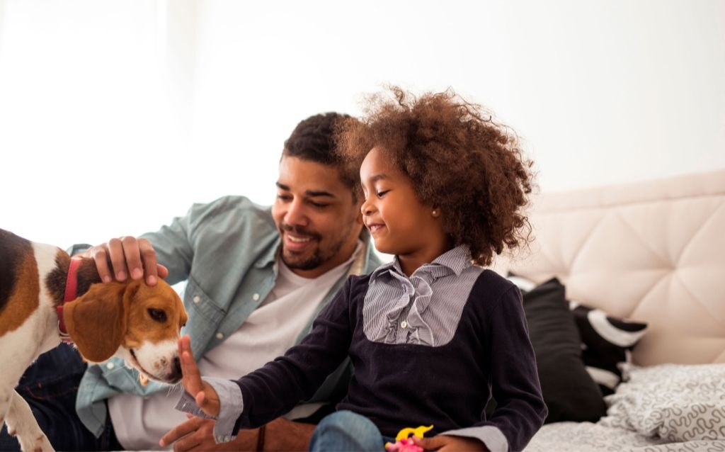 feriados de outubro 2023 pai com uma criança, ambos com a pele negra, sendo que a criança tem um longo cabelo crespo e o pai cabelo curto e cavanhaque. os dois estão brincando com um cachorro beagle
