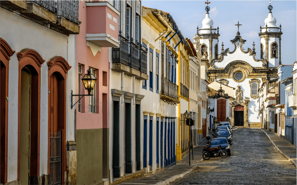 Foto de uma rua de pedras com construções coloridas e tradicionais pelas calçadas. Centralizada ao fundo tem uma igreja.