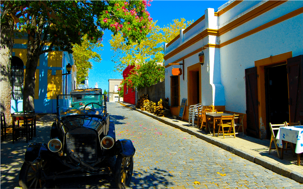 Imagem de um carro antigo em uma rua de pedra em Sacramento. Ao fundo, várias árvores floridas e um céu azul.