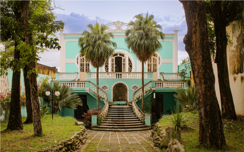 Imagem de um casarão verde com uma escada e duas palmeiras na entrada.
